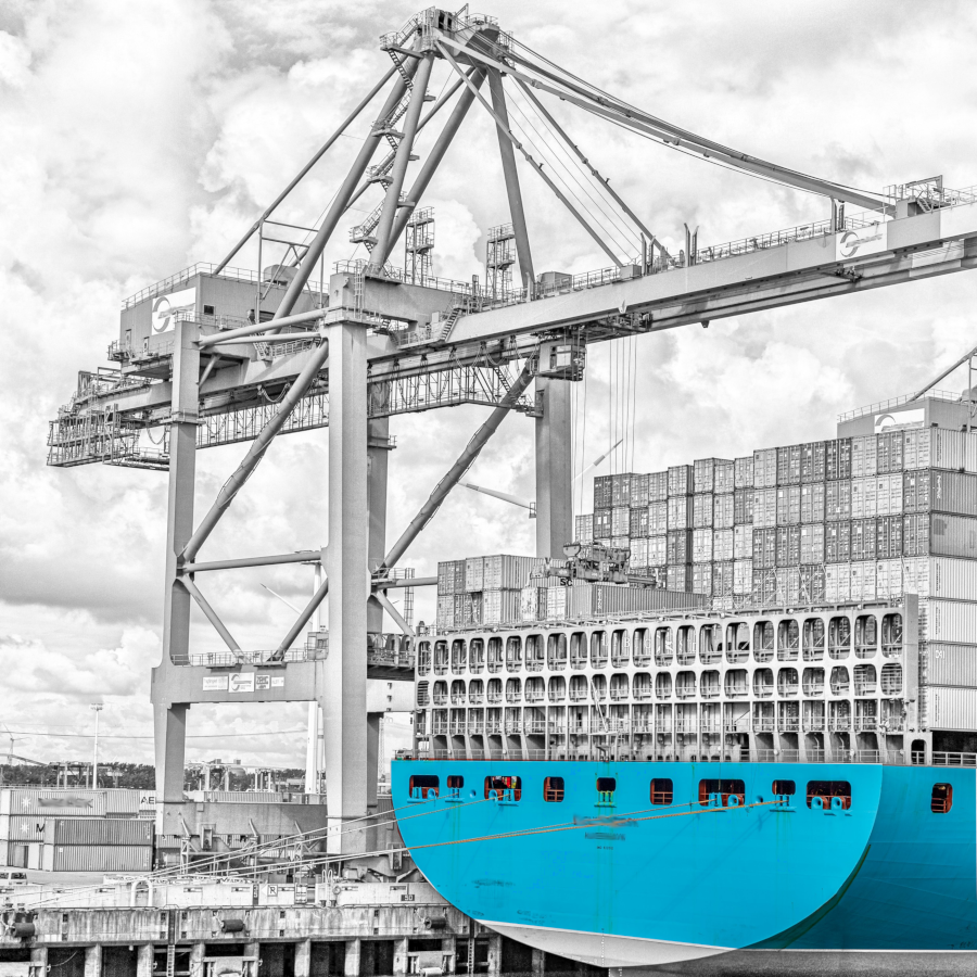 Ein schwarz-weiß Bild eines Frachthafens mit einem Lastkran und einem Schiff, dessen Bug blau gefärbt ist.